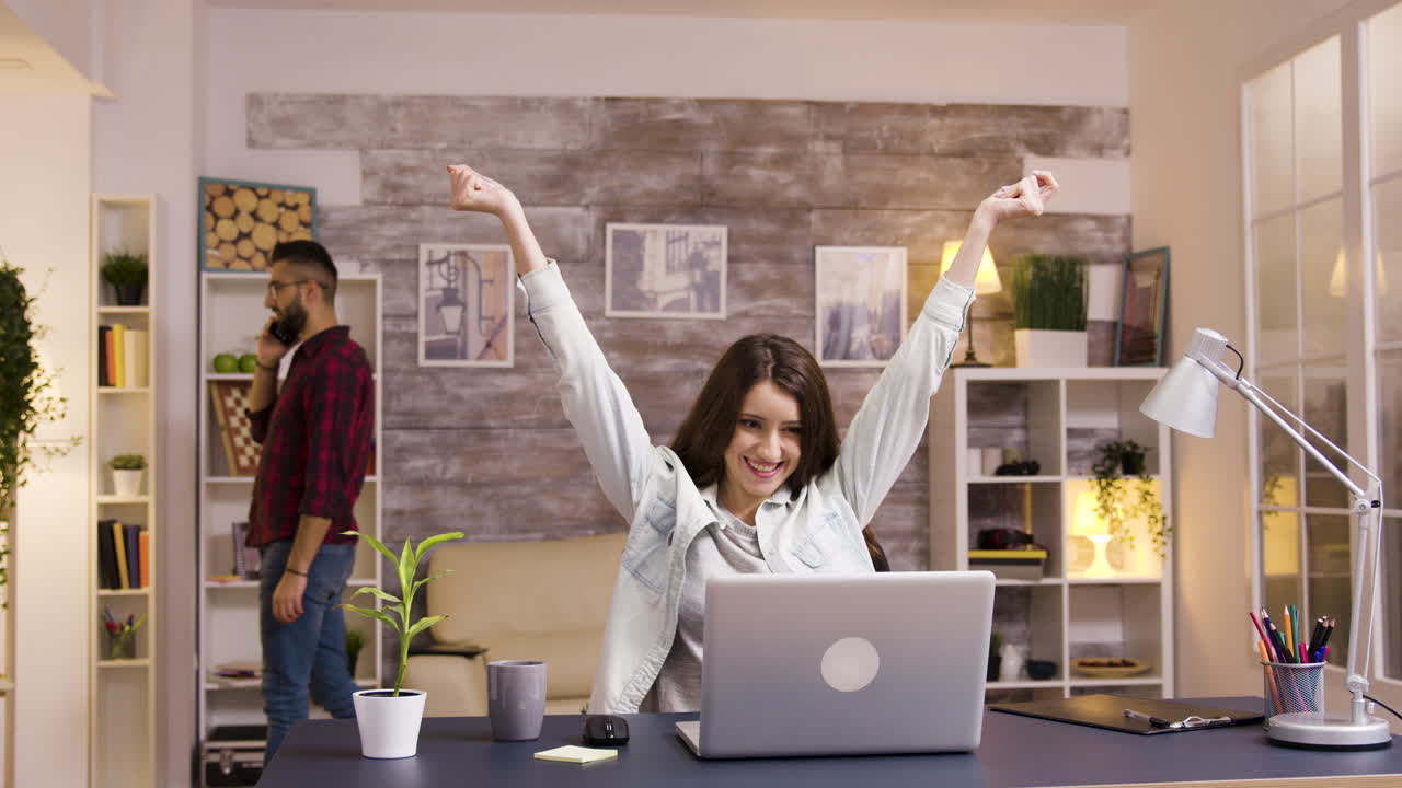 Woman celebrates success in home office with man on phone in background