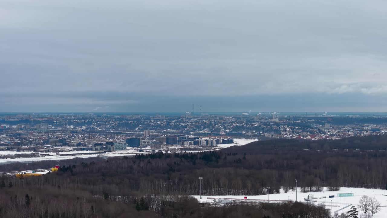 Luftoptagelser, der fanger et stort bylandskab sidestillet med en sneklædt skov. Vidvinkeludsigten fremhæver urbane og naturlige elementer under en overskyet himmel, hvilket skaber en fredfyldt vinterstemning