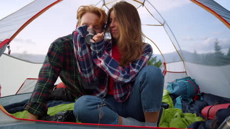 Young-couple-sitting-in-tent-and-drinking-hot-tea-during-hike