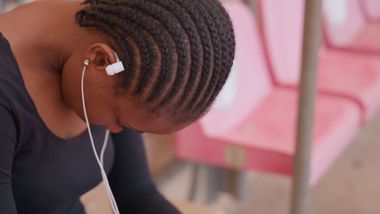 black teen commuter bowed head earbuds showing quiet fatigue and reflection on pink transit seats closeup of cornrow hairstyle and hand touching forehead, candid portrait capturing waiting, mood