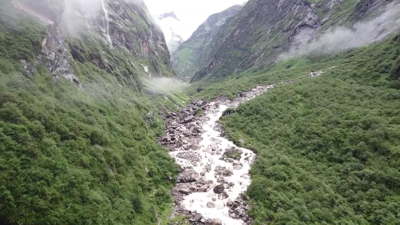 Luftoptagelser, der flyver over en flod i Himalaya, omgivet af barske, majestætiske landskaber med tårnhøje bjerge og frodigt grønt.