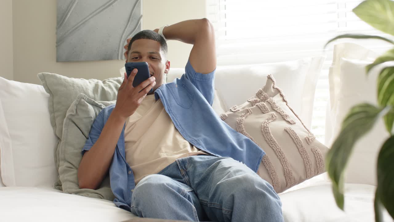 Uomo afroamericano che guarda lo smartphone sul divano del soggiorno, prendendolo per un messaggio vocale