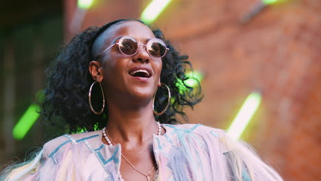 Young-black-woman-wearing-multicoloured-fringed-jacket-dancing-in-the-street-and-laughing,-waist-up,-low-angle