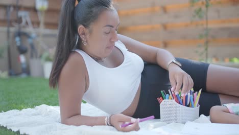 una madre y su hija pequeña se sientan en una manta en el patio trasero, participando en una actividad de dibujo juntos. la escena al aire libre captura un momento familiar pacífico, fomentando la creatividad y el vínculo.