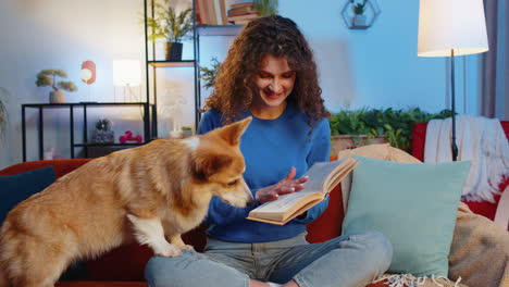 happy woman reading book while sitting beside cute corgi pet dog on sofa in living room at home