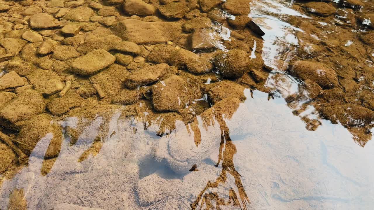 Agua clara y poco profunda de río fluyendo suavemente sobre piedras lisas y un lecho arenoso, suaves reflejos en la superficie, texturas naturales visibles bajo las ondulaciones, escena diurna tranquila.