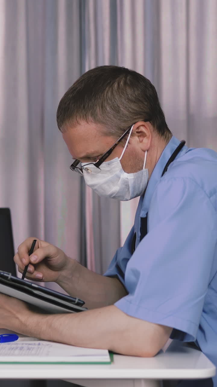 young doctor with glasses and protective mask, sitting at table in his office, where there is laptop, cell, something writes by stylus in medicine tablet