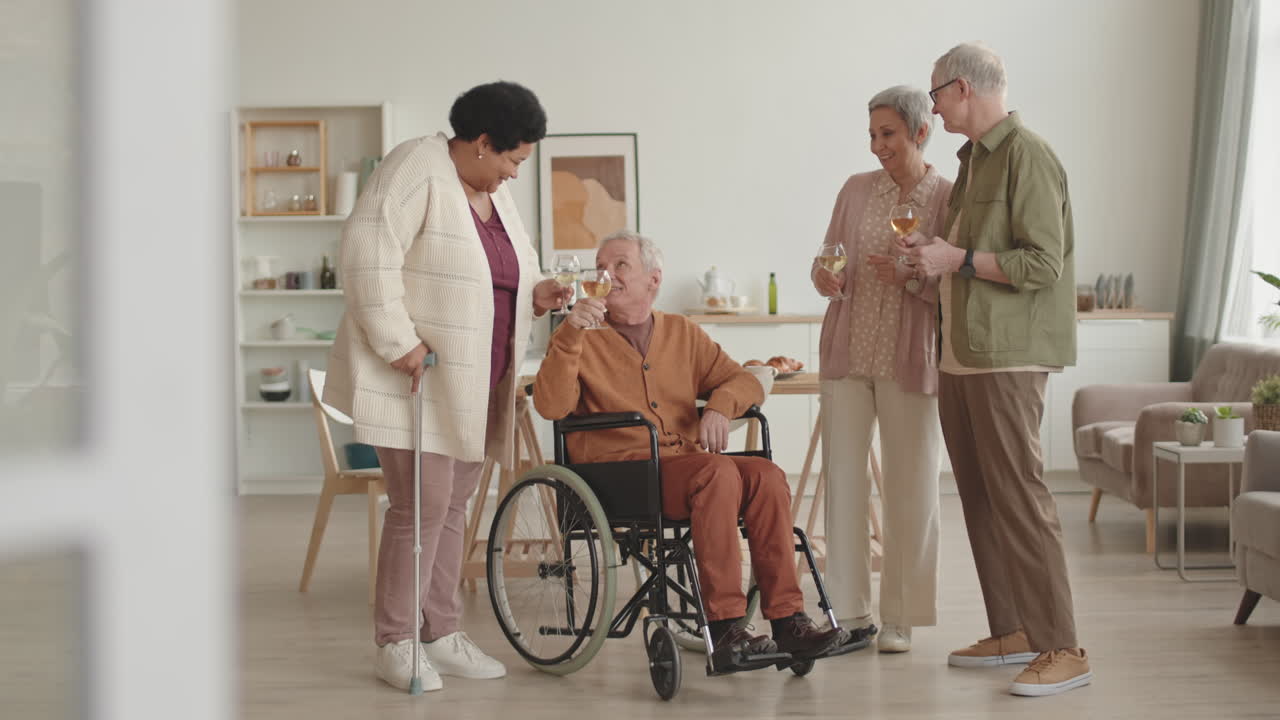 Full shot of African woman leaning on cane, handicapped man in wheelchair, grey-haired Asian lady and senior Caucasian man standing in apartment, drinking white wine
