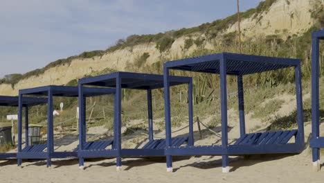 Empty-blue-wooden-beach-tents-with-a-cliff-in-the-distance-at-Fonte-da-Telha-beach-in-Portugal