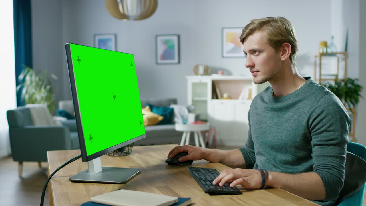 un bel giovane lavora su un computer personale con uno schermo verde mentre è seduto alla sua scrivania nell'accogliente appartamento.