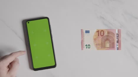 Overhead-Währungsaufnahme-Einer-10-Euro-Note-Neben-Einer-Person,-Die-Ein-Green-Screen-Handy-Verwendet