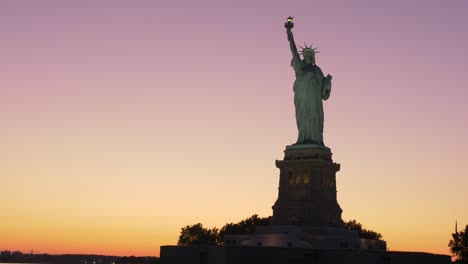 Toma-De-Establecimiento-De-La-Silouhette-De-La-Estatua-De-La-Libertad-Al-Amanecer,-Tomada-Desde-Un-Bote-En-Una-Rampa-Verde-Azulado-Y-Naranja