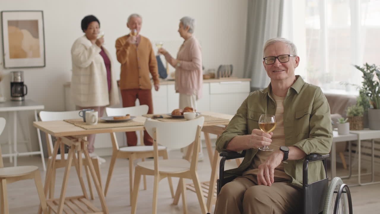 Medium long POV of happy elderly handicapped Caucasian man in wheelchair at home, smiling, raising white wine glass and looking on camera