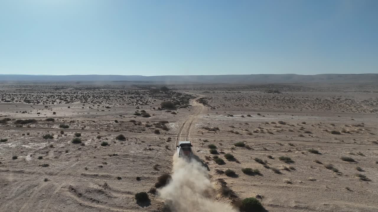 Drone tracking shot of a white off-road vehicle speeding through a vast arid desert landscape, kicking up a massive cloud of sand and dust