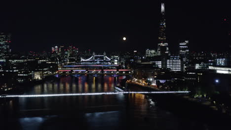 Fly-above-River-Thames-at-Millennium-Footbridge.-Aerial-view-of-illuminated-buildings-and-landmark-at-night.-London,-UK