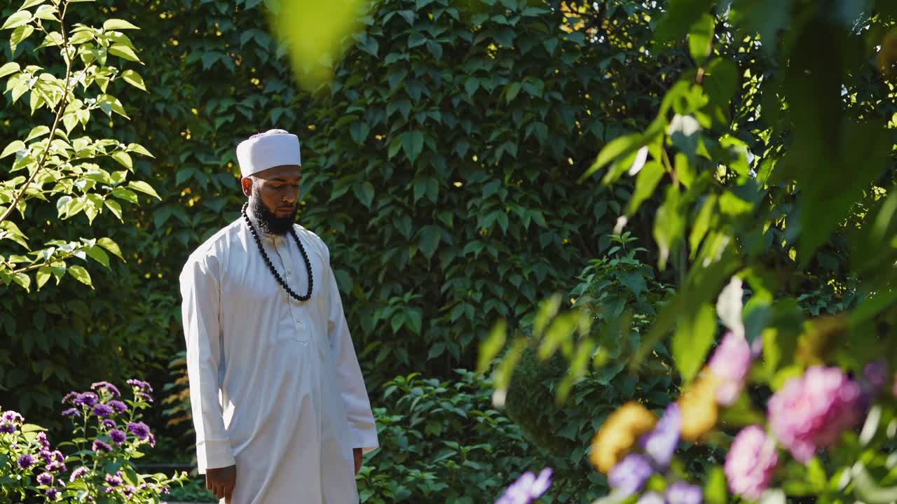 A serene video scene of a man in white traditional attire praying in a lush garden