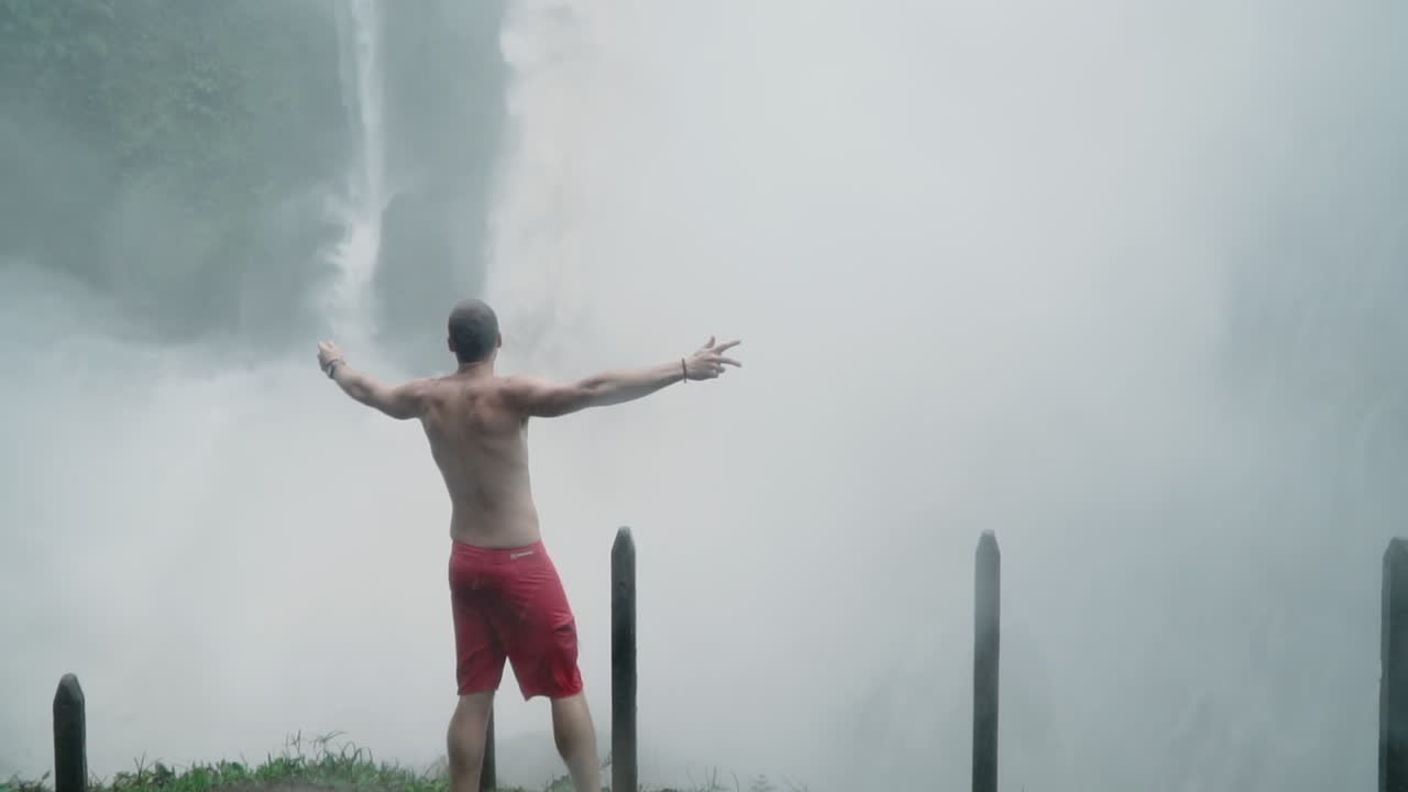 Man celebrates in front of roaring jungle waterfall in pouring rain, slow motion