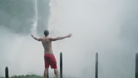 man celebrates in front of roaring jungle waterfall in pouring rain, slow motion