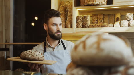 Portrait-Shot-Of-The-Young-Male-Baker-Looking-At-The-Side-While-Holding-A-Tray-With-Bread-Then-Looking-At-The-Camera-In-The-Shop