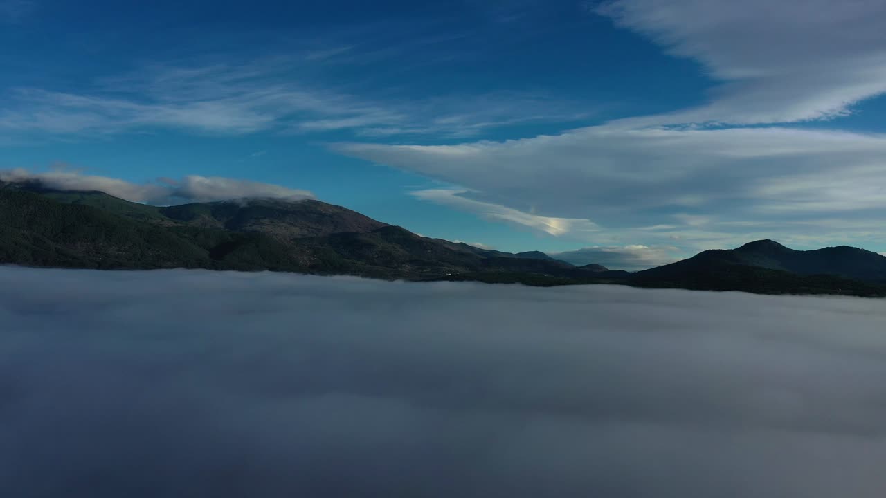 Volo di drone sopra le nuvole, che mostra le montagne più alte con un mare di nuvole a fare da sfondo, sotto un cielo blu con nuvole alte, al mattino d'inverno in una valle ad Avila, Spagna