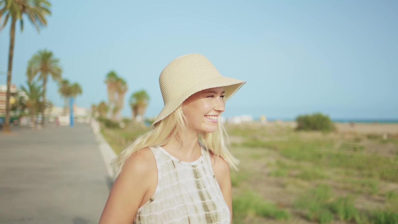 Lachende jonge blonde vrouw met een strohoed op een strandpromenade