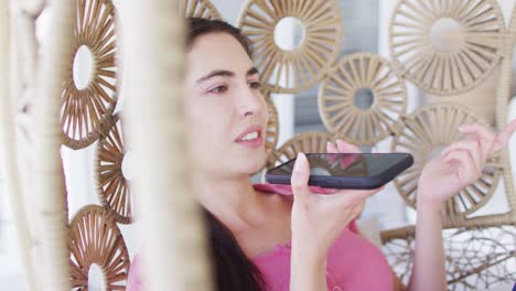 happy caucasian woman sitting on hanging chair on balcony and talking on smartphone