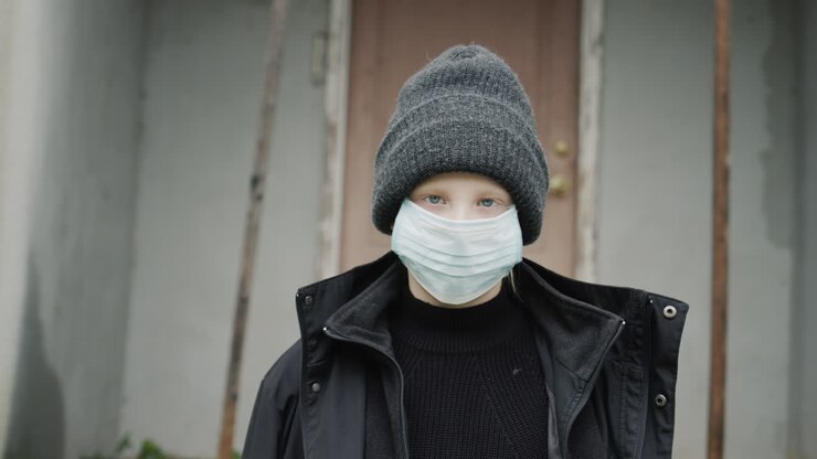 Portrait of a child from a poor family. Standing on the threshold of his house in a protective mask