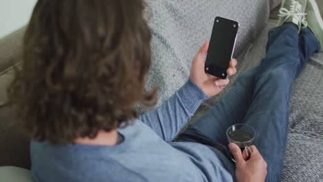 hombre caucásico sentado en el sofá en la sala de estar usando un teléfono inteligente y bebiendo café