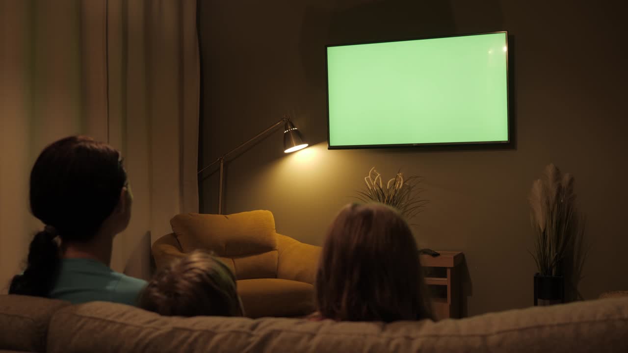 Vista posteriore di una famiglia con bambini seduta sul divano in salotto la sera, che guarda insieme la TV con schermo verde.