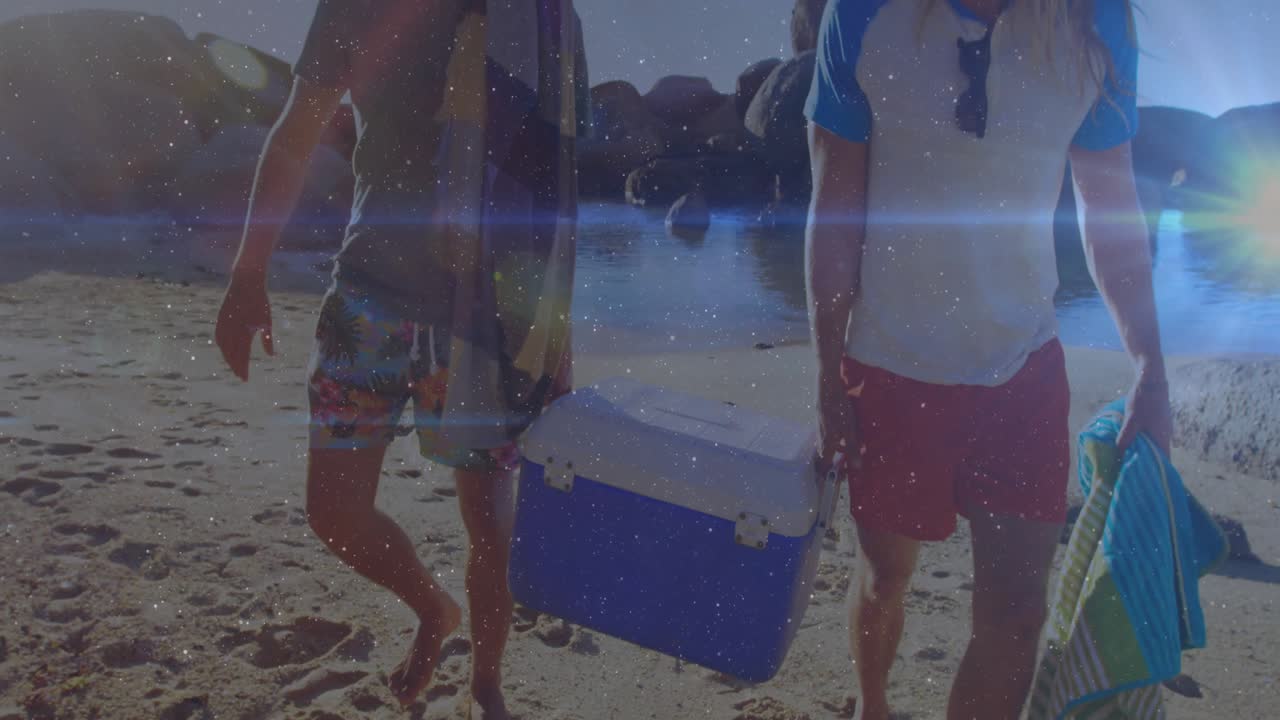 Leisure opening shot capturing two friends walking and carrying cooler toward cove amid lens flares