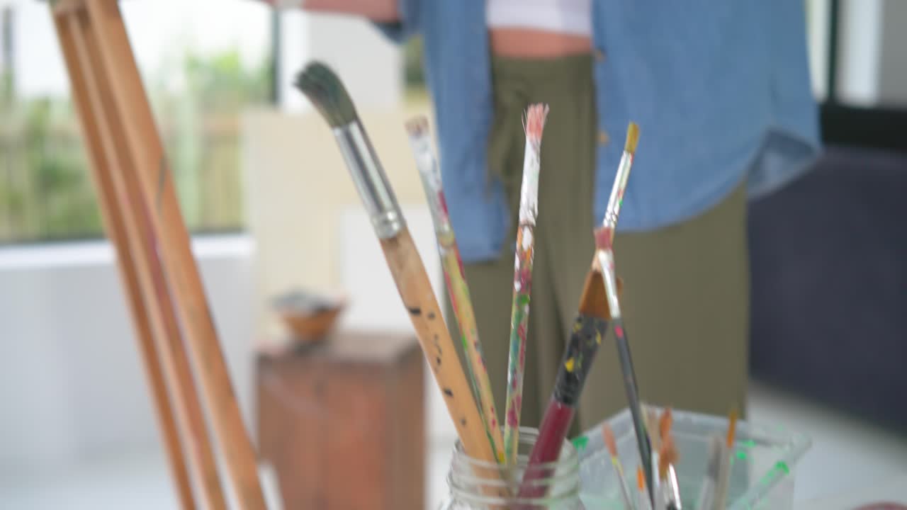 Donna matura che si protende verso la tela, intingendo il pennello, dipingendo per hobby su un cavalletto a casa, con spazio per il testo.