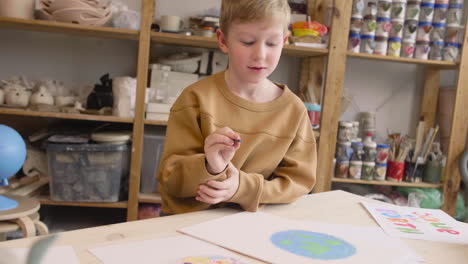 blond kid painting a earth on a paper sitting at a table in a craft workshop where are signs with environmental quotes 2