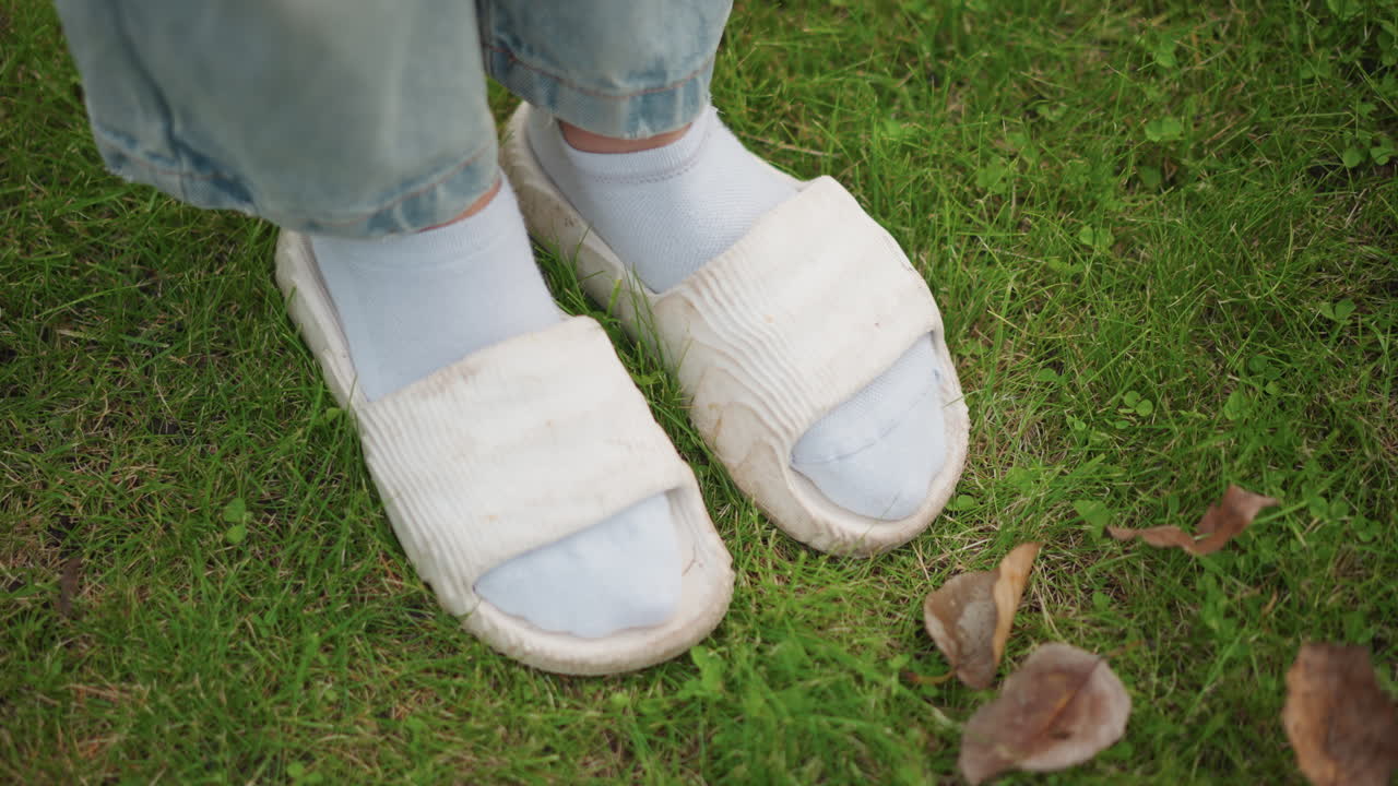 Witte pantoffels en sokken op het gras, oudere voeten naast een tuinvork die bruine bladeren opruimt. Close-up van de geweven textuur van de pantoffel, denim manchetten, lichte tuinklus, rustige huiselijke herfstroutine met tastbare details.