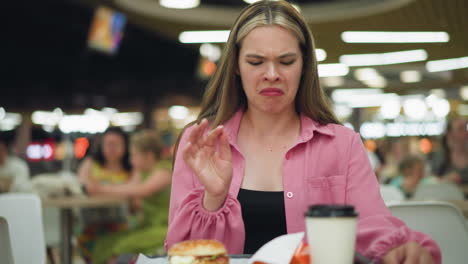 mujer sentada en un restaurante toma una patata frita, la muerde, luego le disgusta el sabor y la lanza hacia abajo, se limpia las manos con una servilleta, mirando contemplativa, con una vista borrosa de la gente a su alrededor