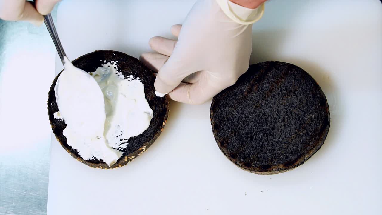 Cook puts white sauce on black burger bun with spoon. Black bread with sauce. Healthy vegan burger. Healthy eating