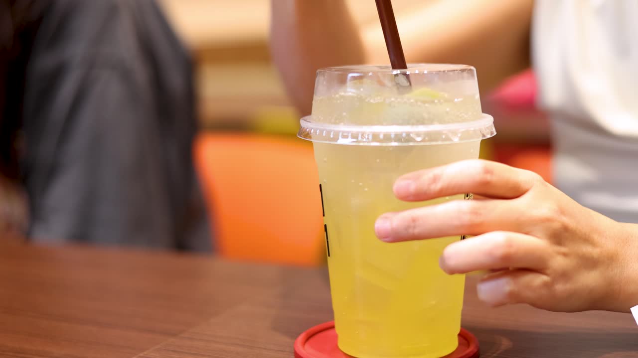 A person stirs a plastic cup of lemon soda with a straw on a wooden table in a brightly lit indoor café, using a close-up perspective