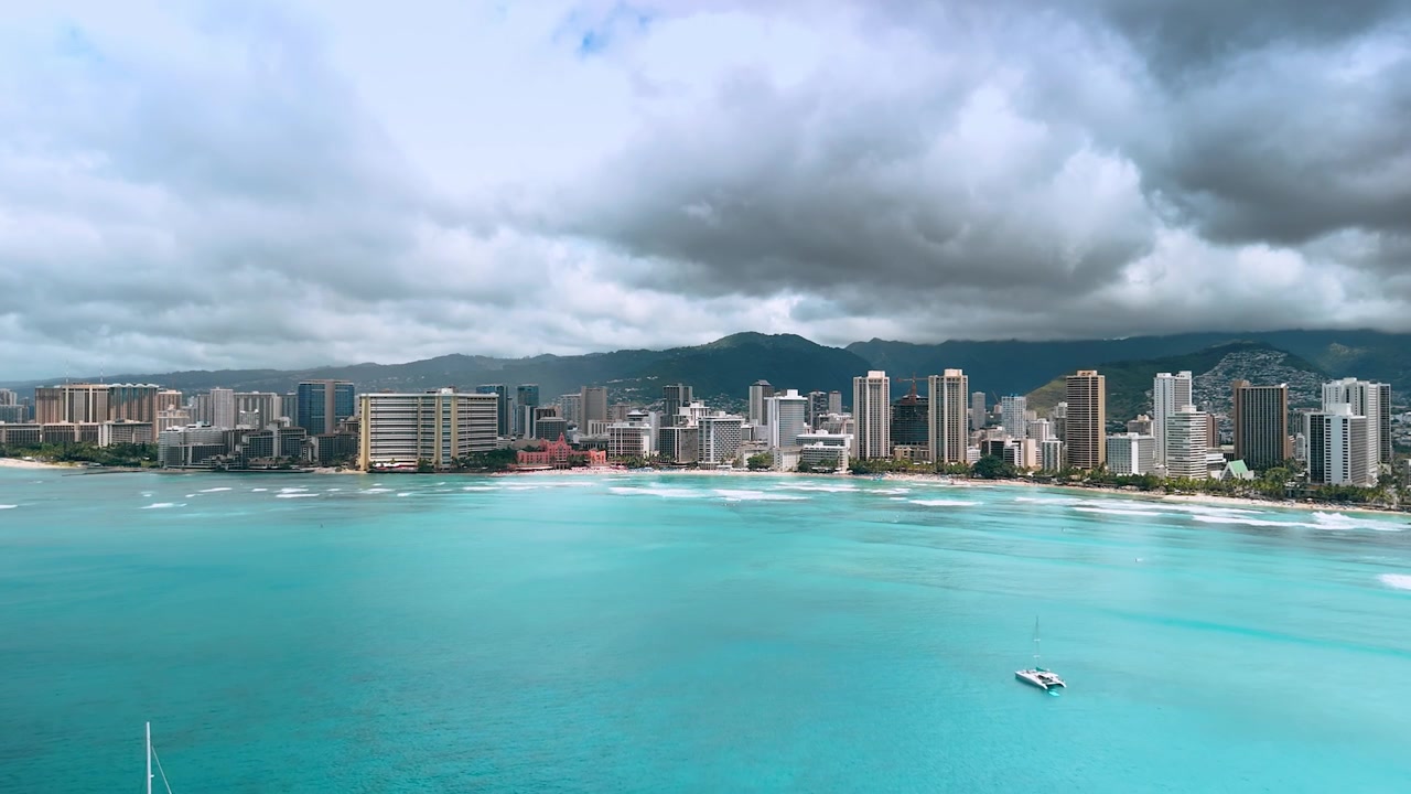 Pesanti nuvole grigie si spostano verso il calmo paesaggio marino turchese dell'Oceano Pacifico. Riprese con drone in avvicinamento allo skyline di Waikiki a Honolulu, Hawaii, USA in una giornata nuvolosa
