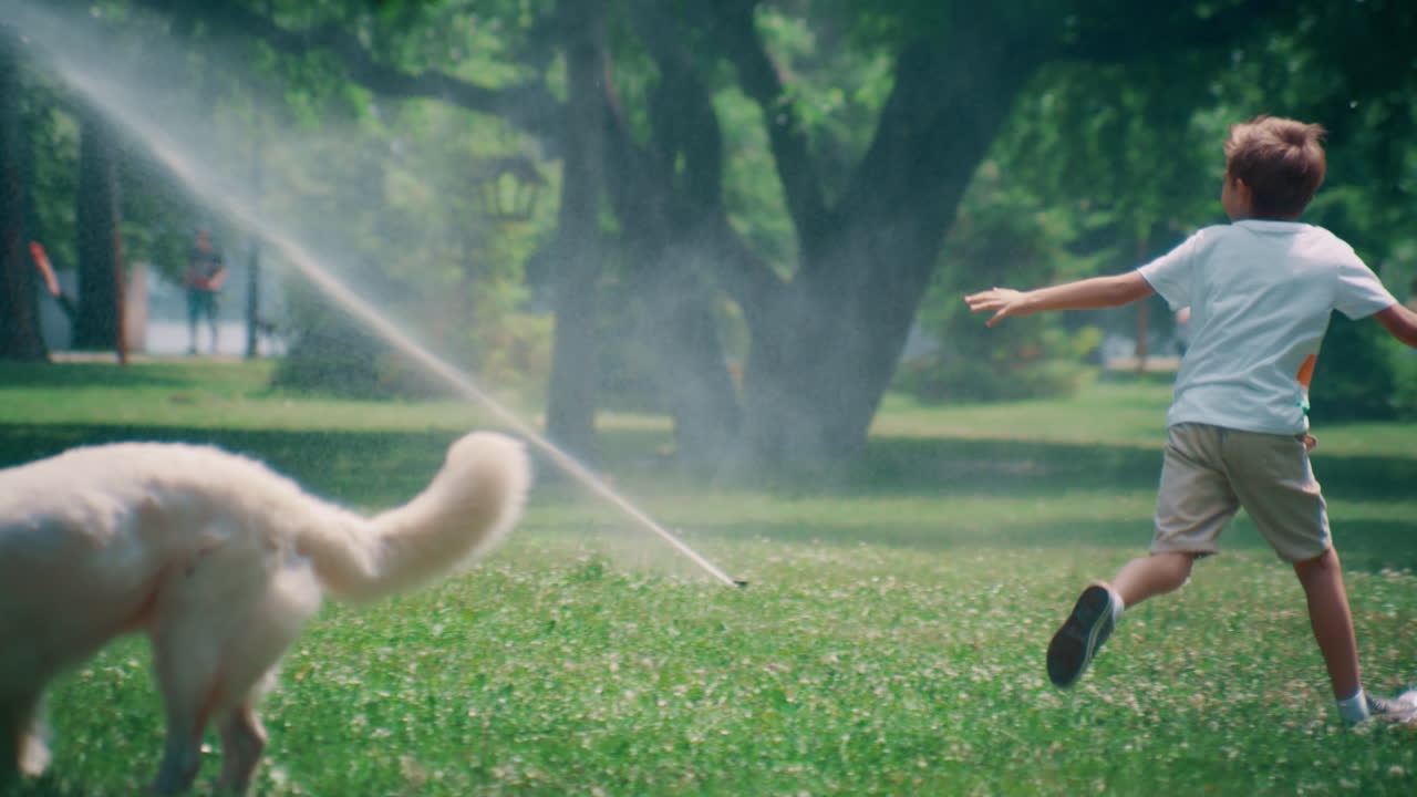 Cheerful little kid running from adorable pet at water sprinklers in summer park