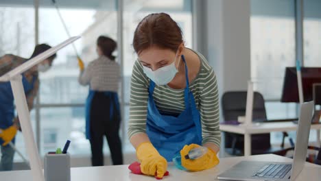 portrait of young female worker in uniform and safety mask cleaning desk with rag and spray in office