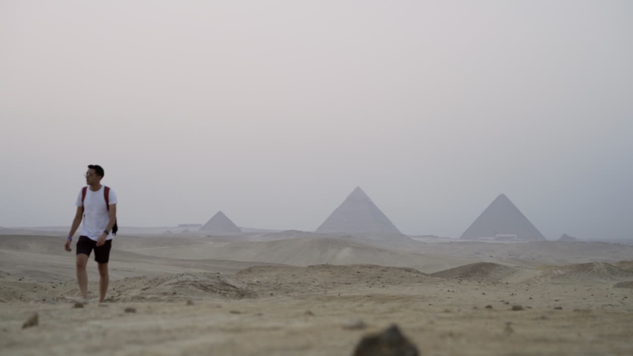 Young Egyptian man walks in desert with Giza pyramids in distance, slow motion