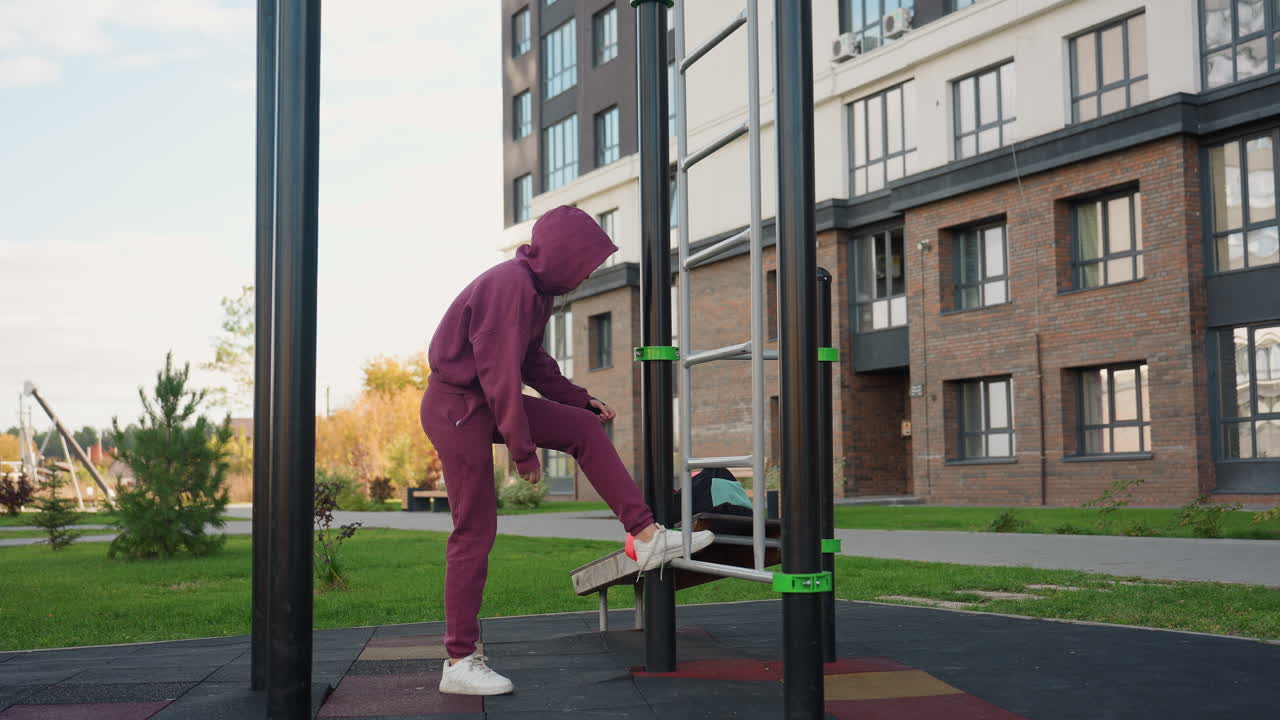 Outdoor trainer in hoodie carrying exercise bag approaches urban calisthenics bars sets bag at corner then bends down to tie shoelace before starting intense workout amid modern building backdrop