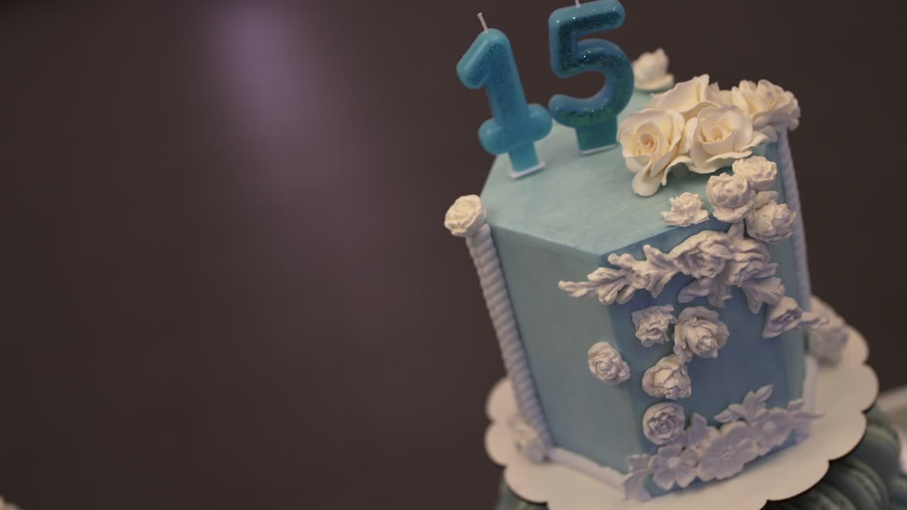 Slow reveal showcases a hexagonal blue fondant cake adorned with cascading white flowers and number candles “15,” highlighting artistic detail and celebration mood for a milestone birthday
