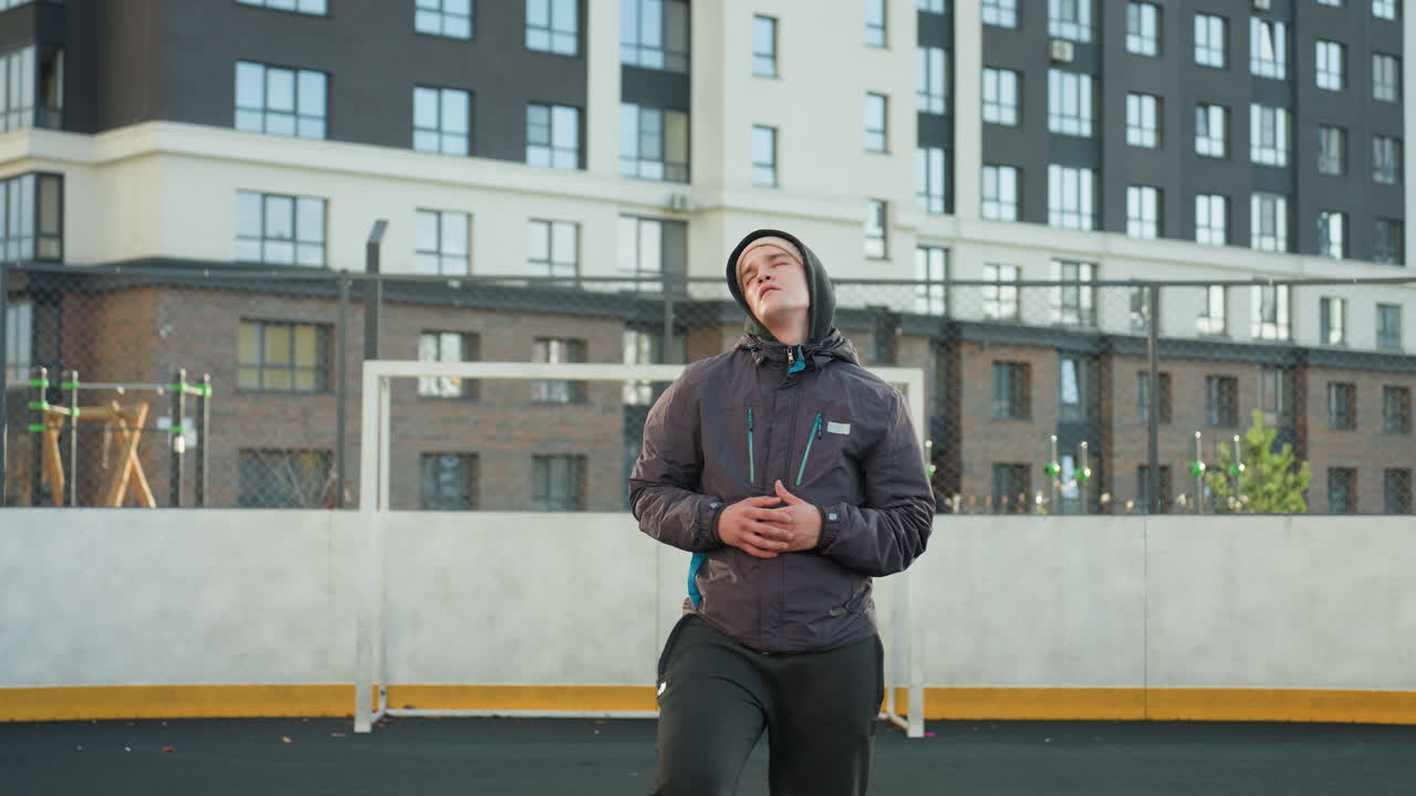 스포츠 선수는 공을 땅에 떨어뜨리고, 자신감 있게 공에 발을 고, 손을 잡고, 고층 주거 건물과 스포츠 인프라로 둘러싸인 현대 도시 스포츠 경기장에서 머리를 돌립니다.