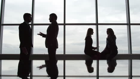 a business meeting between two men on one side and two women on the other side backlit by a large window