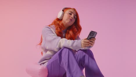 studio shot of young gen z woman wearing headphones streaming music to mobile phone 2