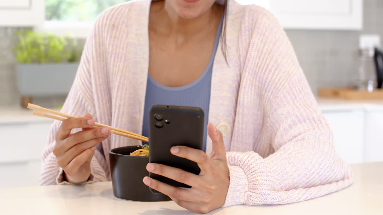 Eating noodles with chopsticks while browsing smartphone at home kitchen