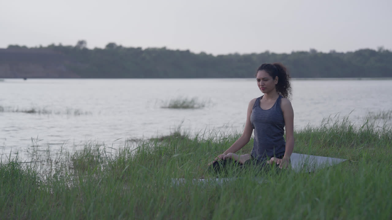 A slow-motion of a South Asian Woman in Lotus Pose (Padmasana) with prayer hands meditating by a lake on a mat