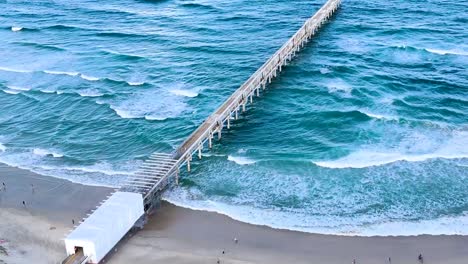 drone footage shows a long pier stretching over vibrant ocean waves and sandy beach.