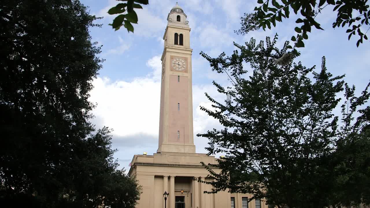 Iconica torre dell'orologio della Memorial Tower nel campus della Louisiana State University (LSU) a Baton Rouge, Louisiana, USA, architettura neoclassica con alberi in primo piano sotto un cielo blu.