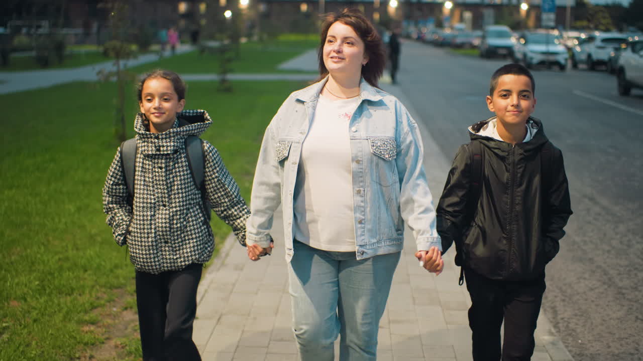 Woman joyfully walks hand in hand with two children along city sidewalk during evening, surrounded by green grass and parked cars, expressing warmth, happiness, and strong family bond in relaxed urban atmosphere
